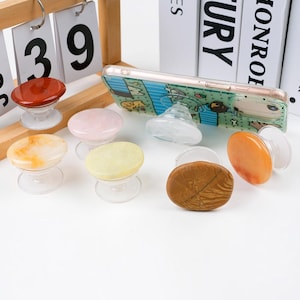 May include: A collection of colorful, gemstone-topped phone grips. The grips are in various colors, including red, pink, yellow, and orange. A phone with a green case and a matching grip is also present. The grips are on a white surface.