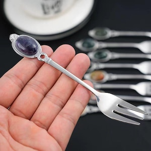 Puede incluir: Un tenedor plateado con un mango decorativo con una piedra preciosa ovalada de color púrpura. El tenedor tiene tres púas y está sostenido en la mano de una persona. Otros tenedores similares con piedras preciosas de diferentes colores son visibles en el fondo.