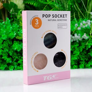 Könnte beinhalten: Eine Schachtel mit drei PopSockets aus Naturstein. Die Schachtel ist weiß und rosa, mit den Aufschriften "POP SOCKET" und "NATURAL GEMSTONE". Die PopSockets sind in Schwarz, Rosa und Blau gehalten.