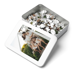 May include: A white puzzle tin with a lid featuring a family photo. The tin is open, revealing a pile of puzzle pieces. The lid displays a color image of a family of five. The puzzle pieces are a mix of white, brown, and green.