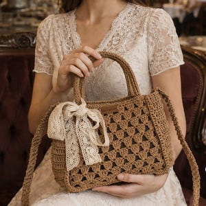 May include: A brown crochet handbag with a rectangular shape and a cream lace bow. The bag features a handle and a long strap. The crochet pattern is visible throughout the bag's design, offering a textured appearance.
