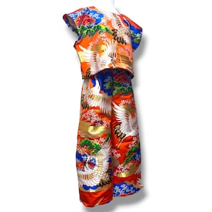 May include: A two-piece outfit consisting of a short-sleeved top and wide-leg trousers. The fabric is a vibrant orange featuring a repeating pattern of white cranes, green pine trees, blue clouds, and pink cherry blossoms with gold accents throughout.