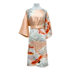 May include: Peach-coloured kimono-style robe with wide sleeves and a matching belt. The robe features a print of red and white cranes in flight against a cloudy background. The robe is displayed on a mannequin.