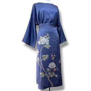 Puede incluir: Un vestido azul marino de largo midi con mangas anchas y sueltas y una cintura ajustada. La falda muestra un estampado botánico detallado de peonías blancas y rosa claro con hojas verdes y ramas finas marrones desde el dobladillo.