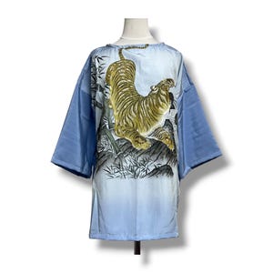 May include: A light blue kimono-style top with a large illustration of a golden tiger on a rocky landscape. The sleeves are a darker shade of blue. The garment is displayed on a mannequin.