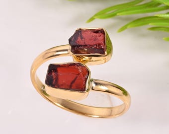 May include: A gold ring with two raw, rectangular, deep red gemstones. The ring's band curves around the finger, with the gemstones set in a bezel setting. The background is white with green foliage.