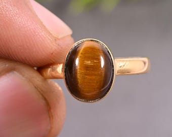 May include: A gold-toned ring featuring a smooth, oval-shaped tiger's eye gemstone. The stone displays a characteristic chatoyant brown and golden luster. The metal band is plain and polished, holding the cabochon-cut stone in a simple bezel setting.