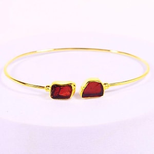 Könnte beinhalten: Ein goldfarbenes Armreif-Armband mit zwei rechteckigen, roh geschnittenen, roten Edelsteinen. Das Armband hat ein schlichtes, offenes Design, wobei die Edelsteine an den Enden des Bandes eingesetzt sind.