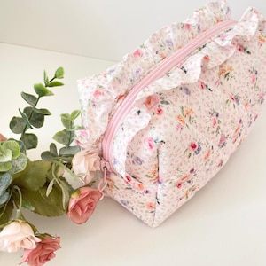Könnte beinhalten: Eine Kosmetiktasche mit floralem Muster, rosa Reißverschluss und Rüschenbesatz. Die Tasche hat ein Muster aus kleinen Blumen in Rosa-, Lila- und Gelbtönen auf weißem Grund. Die Tasche ist rechteckig.