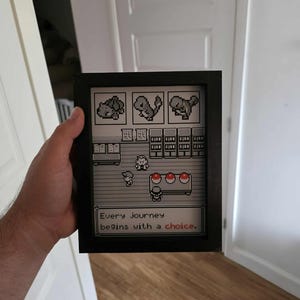 May include: Black framed pixel art featuring a scene from a video game. The artwork depicts a retro game scene with characters, items, and the text "Every journey begins with a choice."