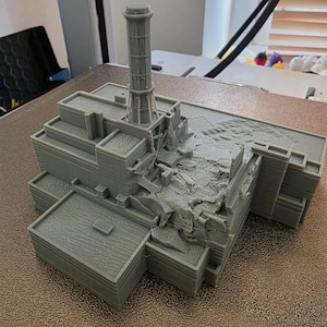 Op de afbeelding: Een grijs 3D-geprint model van een gebouw met een hoge cilindrische structuur en een beschadigd gedeelte. Het model heeft een gedetailleerd architectonisch ontwerp, met meerdere niveaus en een getextureerd oppervlak. Het thema is een miniatuur architectuurmodel.