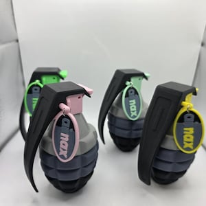 May include: Four novelty grenade-shaped objects. Each has a black handle and a coloured pin, with a matching tag reading "dinax". The grenades are mainly grey, black, and blue, with pink, green, and yellow accents. The background is white.