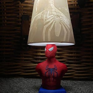 Può includere: Lampada di Spider-Man rossa e blu con paralume beige. Il paralume presenta un'immagine dettagliata della schiena di Spider-Man. La base della lampada è blu e riporta la scritta "SPIDER-MAN".