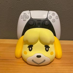 Op de afbeelding: Een gele Isabelle-figuur, met zwarte ogen en een lachende mond, staat voor een wit-zwarte PlayStation 5-controller. De controller heeft een zwart touchpad en knoppen. De figuur is van Animal Crossing.