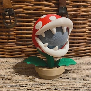 Może przedstawiać: Głośnik w kształcie czerwonej i białej rośliny Piranha z serii gier wideo Super Mario. Głośnik ma czerwoną głowę z białymi kropkami, białe zęby i zielone liście. Jest umieszczony w beżowej doniczce.
