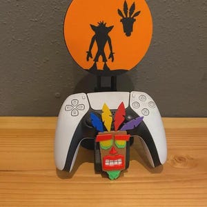 Op de afbeelding: Een witte en zwarte PlayStation 5-controller met een kleurrijk Aku Aku-masker en een silhouet van Crash Bandicoot tegen een oranje achtergrond. De controller rust op een houten oppervlak, met een donkergrijze muur op de achtergrond.