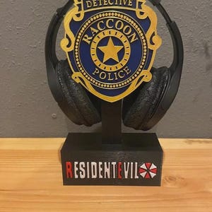 Può includere: Supporto per cuffie nero con distintivo blu e oro della Raccoon Police, con la scritta "Detective". La base mostra "Resident Evil" in rosso e bianco. Accessorio per gamer.