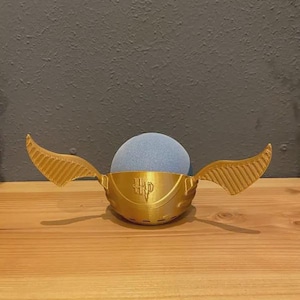 May include: A golden-colored, 3D-printed Snitch from the Harry Potter series. The Snitch has two wings and a round body, with the letters "HP" on the front. A blue sphere is inside the Snitch.