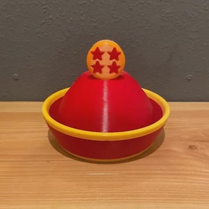 Puede incluir: Un objeto rojo y amarillo en forma de cuenco con una cúpula roja en el centro. Encima de la cúpula hay una pequeña esfera naranja con cuatro estrellas rojas. El objeto está sobre una superficie de madera.