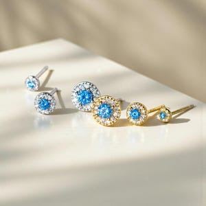 May include: Several pairs of stud earrings in silver and gold. Each earring has a central light blue gemstone encircled by smaller, clear stones. The earrings are in different sizes, including small and larger pairs.