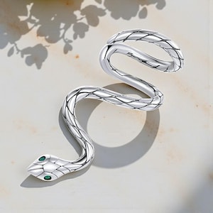 May include: A silver snake ring with a textured pattern. The snake's head has two green gemstone eyes. The ring is coiled in a serpentine shape, creating a unique design. The ring is displayed on a light-coloured surface.