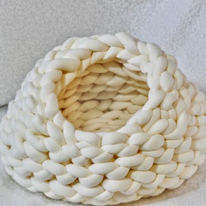 Puede incluir: Una cesta de color crema tejida a mano con forma redonda. La cesta está hecha de hilo grueso y trenzado, creando una superficie texturizada. La parte superior abierta revela el interior, también hecho del mismo hilo. La cesta está sobre una superficie blanca.