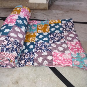 May include: A colorful patchwork quilt, partially rolled and partially unfolded. The quilt features a variety of patterned squares in shades of pink, blue, purple, and yellow. The patterns include floral and leaf designs.