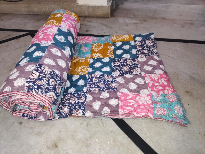 May include: A colorful patchwork quilt, partially rolled and partially unfolded. The quilt features a variety of patterned squares in shades of pink, blue, purple, and yellow. The patterns include floral and leaf designs.