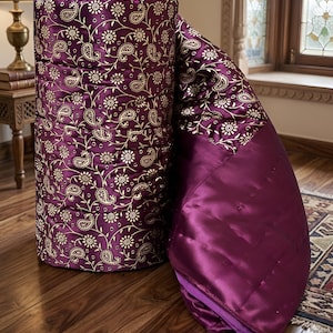 Könnte beinhalten: Eine aufgerollte, gesteppte Bettdecke in tiefem Lila, mit einem goldenen Paisley- und Blumenmuster. Die Bettdecke ist teilweise ausgerollt und zeigt einen glatten, satinartigen Stoff in einem passenden Lilaton.