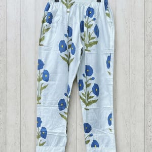 May include: White pajama pants with a blue floral pattern. The pants have a drawstring waist and are made of a lightweight fabric. The blue flowers are stylized and repeated across the fabric, with green leaves and stems.