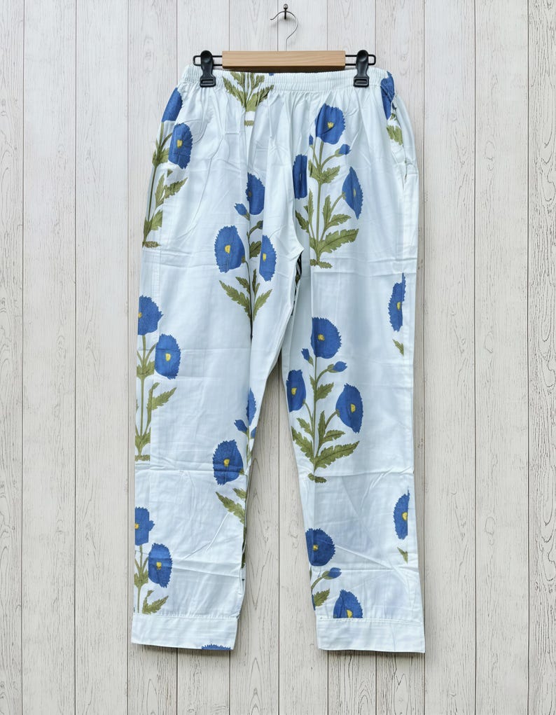 May include: White pajama pants with a blue floral pattern. The pants have a drawstring waist and are made of a lightweight fabric. The blue flowers are stylized and repeated across the fabric, with green leaves and stems.