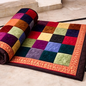 Op de afbeelding: Een opgerolde patchworkquilt met een herhalend vierkant patroon in tinten rood, paars, groen en blauw. De quilt heeft een decoratieve rand met een oranje en gouden ontwerp.