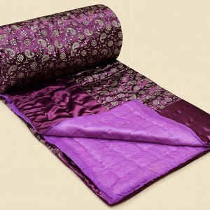 May include: A rolled-up, quilted purple blanket with a paisley pattern in silver and white. The blanket is partially unrolled, revealing a quilted, solid purple interior and a deep purple border. The fabric has a satin sheen.