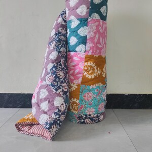 May include: A rolled-up patchwork quilt with a variety of floral and leaf patterns in shades of pink, teal, purple, and gold. The quilt is made of multiple fabric squares sewn together, creating a colorful and textured design.