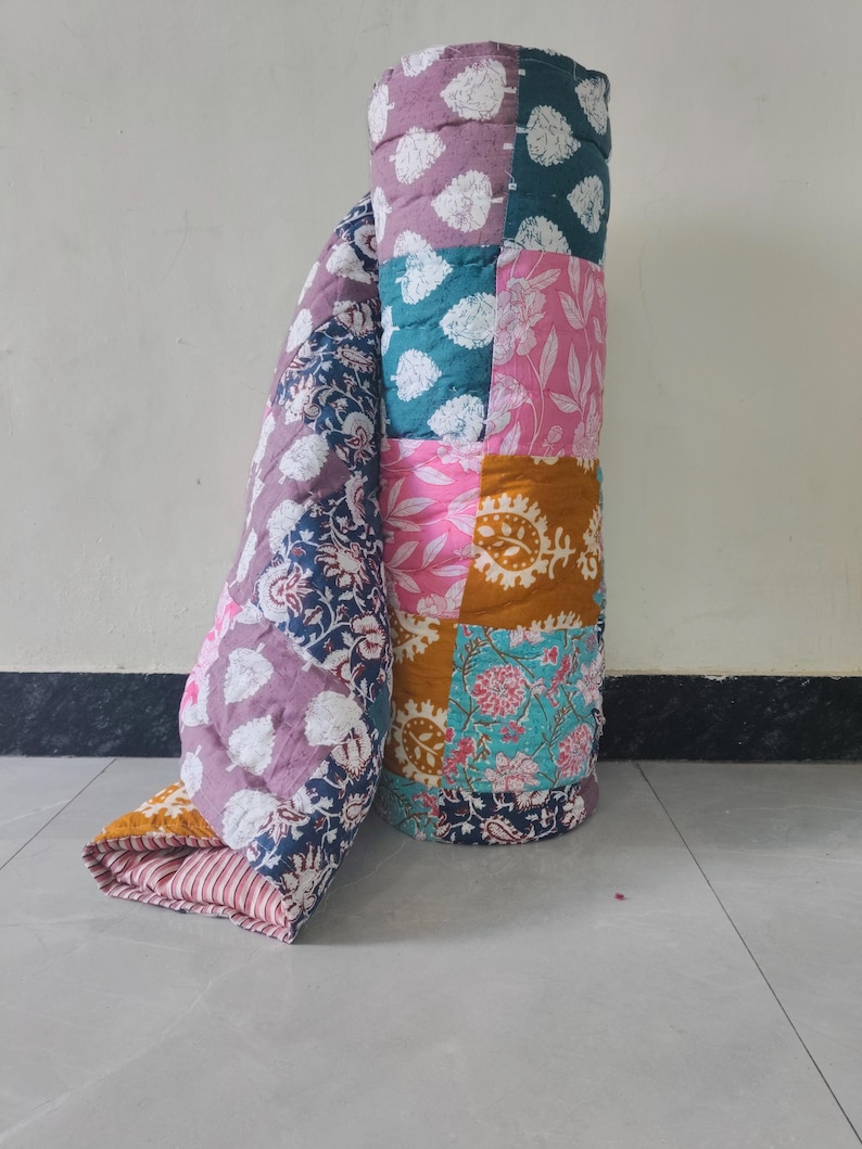 May include: A rolled-up patchwork quilt with a variety of floral and leaf patterns in shades of pink, teal, purple, and gold. The quilt is made of multiple fabric squares sewn together, creating a colorful and textured design.