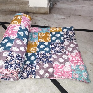 May include: A colorful patchwork quilt with floral and leaf patterns in shades of pink, blue, gold, and purple. The quilt is partially rolled up, revealing its layered construction. The quilt is made of squares of fabric.