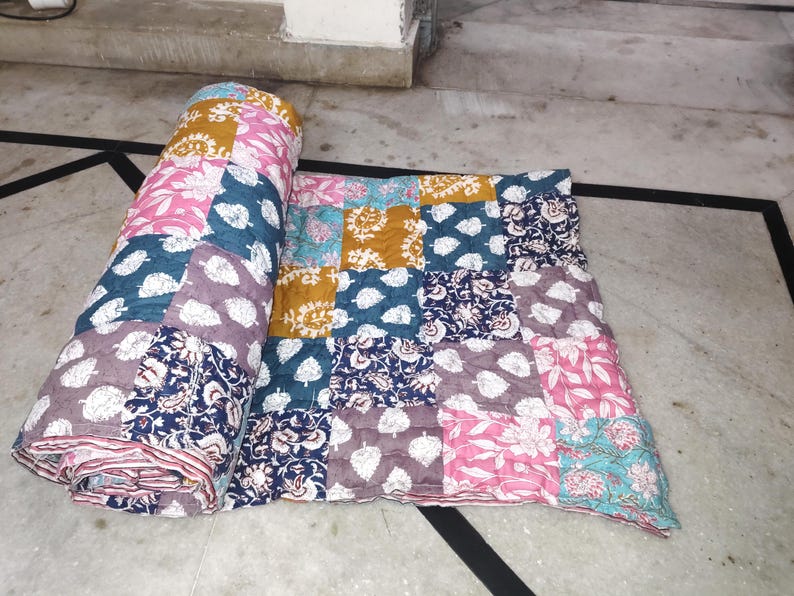 May include: A colorful patchwork quilt with floral and leaf patterns in shades of pink, blue, gold, and purple. The quilt is partially rolled up, revealing its layered construction. The quilt is made of squares of fabric.