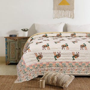 May include: Cream-colored quilt with a repeating pattern of colorful camels. The quilt has a floral border in shades of orange, green, and pink. A matching pillow and a beige mug are on the bed. The quilt is on a bed with white pillows.