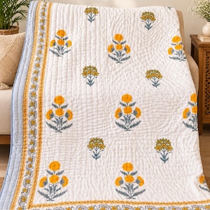 May include: A white quilted blanket featuring a repeating pattern of yellow flowers with gray stems and leaves. The border consists of a yellow floral vine motif framed by thin blue and white striped edges on all sides of the fabric.