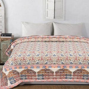 Op de afbeelding: Een sprei met een bloemenpatroon in rood, blauw en oranje tinten. De sprei heeft een decoratieve rand met een boogontwerp. Het bed is opgemaakt met twee witte kussens.