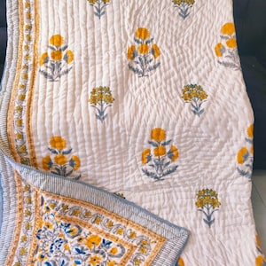 Könnte beinhalten: Eine weiße, gesteppte Decke mit einem sich wiederholenden Muster aus gelben Blumen mit grauen Stielen und Blättern. Die Decke hat einen dekorativen Rand mit gelbem Blumenmuster und feinen blauen Streifen an den Kanten mit detaillierten Nähten.
