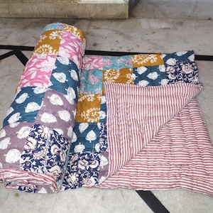 May include: A rolled-up patchwork quilt with a variety of floral and leaf patterns in pink, teal, mustard yellow, and navy blue. The quilt's underside features a red and white striped pattern. The quilt is made of soft fabric.
