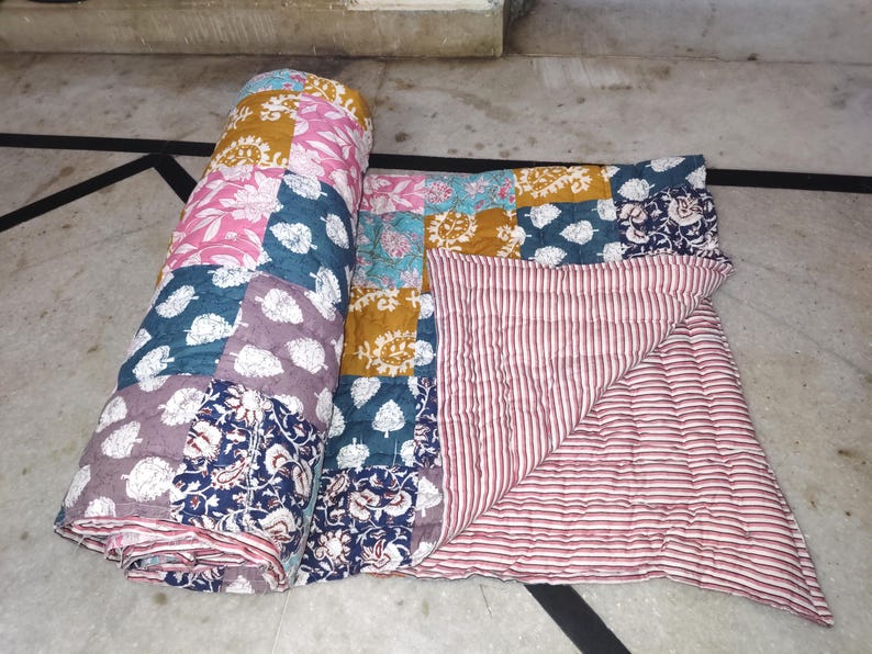 May include: A rolled-up patchwork quilt with a variety of floral and leaf patterns in pink, teal, mustard yellow, and navy blue. The quilt's underside features a red and white striped pattern. The quilt is made of soft fabric.