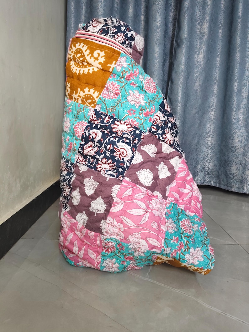 May include: A colorful patchwork quilt with floral and paisley patterns in shades of pink, teal, navy, and mustard. The quilt is folded and appears to be made of cotton fabric. The design features a variety of shapes and sizes.