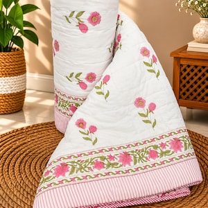 May include: A white quilted blanket featuring a repeating pattern of small pink flowers with green stems. The edges are finished with a wide border displaying a detailed pink and green floral print and a thin pink and white striped trim.