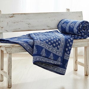 May include: A blue and white patterned quilt, partially rolled and draped over a distressed white bench. The quilt features a detailed floral design. A small potted plant and books are visible in the background.