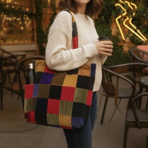 Puede incluir: Un bolso tote patchwork con un colorido patrón de tablero de ajedrez en rojo, amarillo, azul y verde. El bolso tiene una botella de agua negra en el bolsillo lateral. Una persona lleva el bolso, vestida con un suéter color crema y vaqueros azules.
