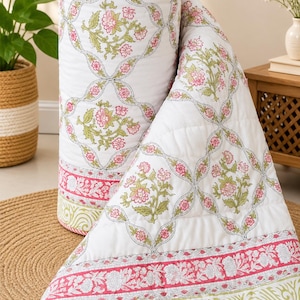 May include: A white quilted blanket featuring a repeating pattern of pink flowers and green leaves enclosed in thin dark outlines. The blanket has a wide border with a dense floral print in pink and white, followed by a light green scroll pattern.