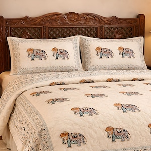 Puede incluir: Juego de cama color crema con un estampado de elefantes repetido en tonos naranja, azul y marrón. La ropa de cama está sobre una estructura de cama de madera con cabecero tallado. Dos lámparas con base dorada están sobre las mesitas de noche.