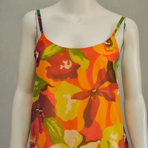 Pode incluir: Um vestido maxi sem mangas com uma estampa floral vibrante. O vestido apresenta um fundo laranja brilhante com flores grandes e coloridas em tons de rosa, verde, amarelo e roxo.